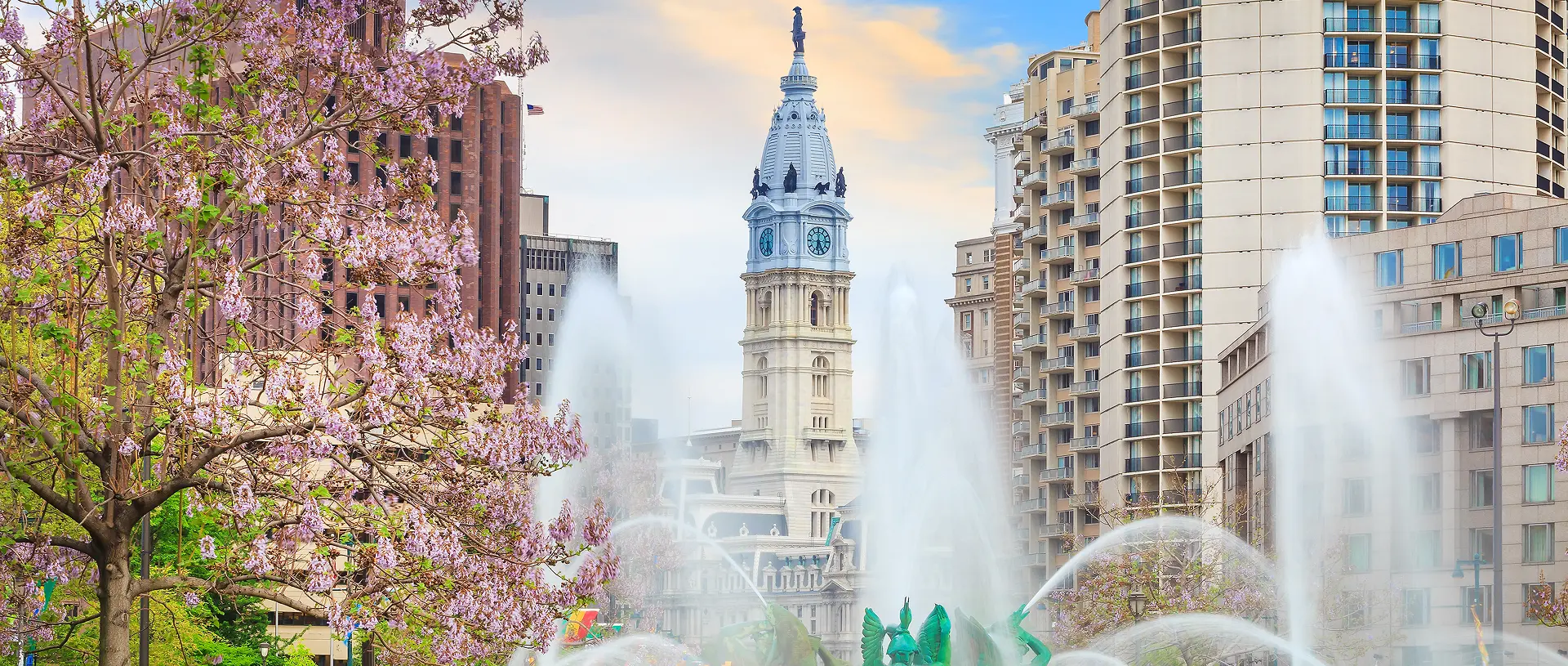 Bustling cityscape with fountains spraying water in the foreground, surrounded by cherry blossom trees. A historic clock tower peaks above modern buildings.