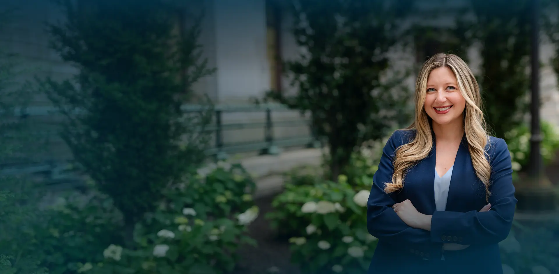 A woman in a blue blazer stands with arms crossed, smiling in front of a garden with green plants and a building in the background.